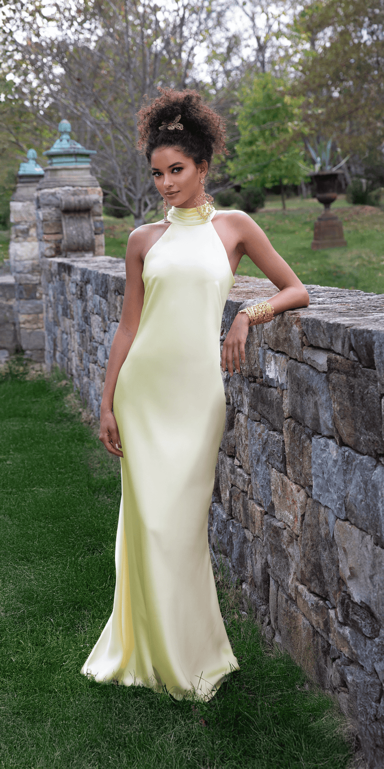 Woman in a light yellow evening gown standing against a stone wall with greenery in the background